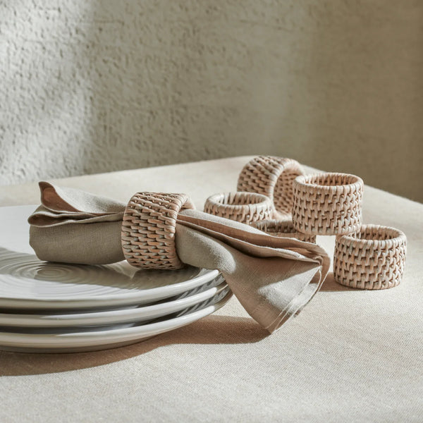 Natural woven tableware set including a napkin ring and matching small holders arranged on a beige tablecloth beside stacked white plates against a textured beige wall.