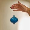 A hand holding a hand-blown glass ornament in a deep teal-blue color with horizontal swirls, featuring a brown hanging stem at the top.