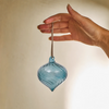 A hand holding a hand-blown glass ornament in a blue color with horizontal swirls, featuring a brown hanging stem at the top.