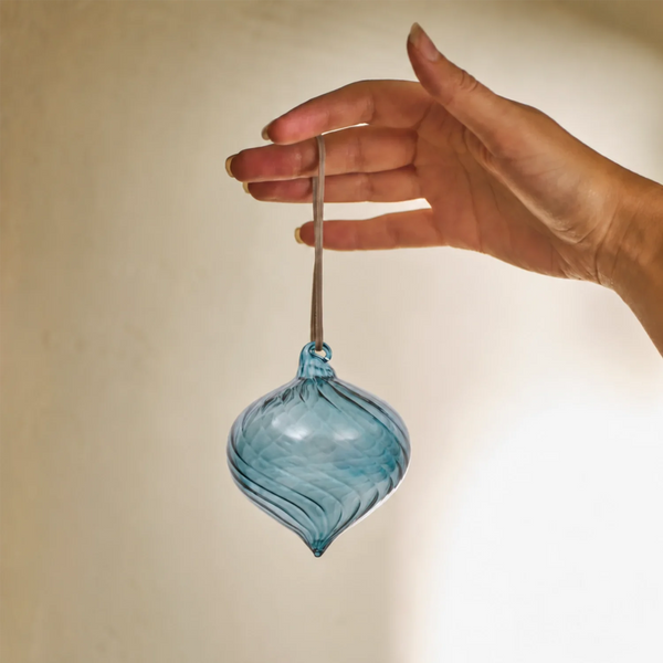 A hand holding a hand-blown glass ornament in a blue color with horizontal swirls, featuring a brown hanging stem at the top.