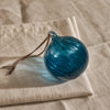 A round, swirled blue glass ornament with a brown hanging string, resting on a textured beige linen cloth.