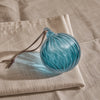 A round, ribbed glass ornament in a translucent light blue color with a brown hanging string, resting on a beige linen cloth.