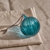 A teal blue ribbed glass ornament with a brown hanging string, resting on a beige linen cloth.