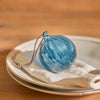 A blue ribbed glass Christmas ornament with a hanging loop, resting on a beige napkin atop stacked white plates on a wooden table.