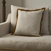 A square throw pillow in a textured cream linen fabric with a contrasting mustard-brown velvet border, styled on a light beige upholstered armchair.