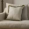 A square off-white linen throw pillow with an olive green velvet piped border, styled on a cream-colored sofa.