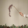 A hand wearing a white long-sleeve shirt holds a decorative branch with clusters of dark red berries against a beige paneled wall, with a garland of pine branches, eucalyptus, and small lights along the bottom.