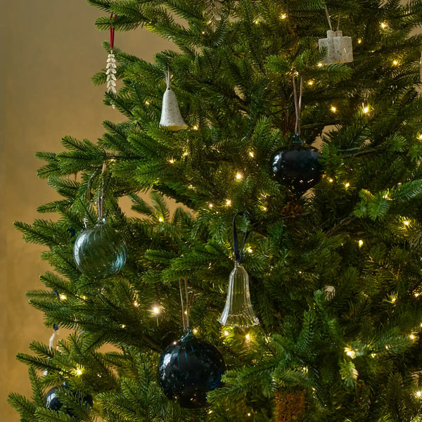 A close-up of a decorated Christmas tree with green branches, warm white string lights, and hanging ornaments including dark blue glass balls, a white bell ornament, and a small white gift box ornament.