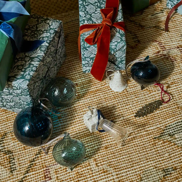 A styled flat-lay of vintage Christmas ornaments and wrapped gifts on a woven beige rug. The ornaments include dark blue glass spheres, a small ribbed clear glass ornament, a white speckled bell-shaped ornament, and a gold pinecone ornament.