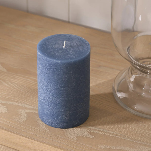 A textured blue pillar candle with a white wick sits on a light wood tabletop next to a clear glass vessel.
