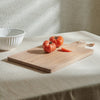 A rectangular wooden cutting board with a hanging loop, styled on a beige linen tablecloth with tomatoes and a carrot slice on top, next to a white ceramic bowl.