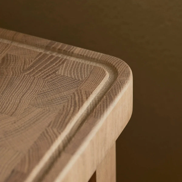 Close-up of Neptune Borough Butchers Block end grain cutting board with rounded corners and a recessed cutting edge, shown against a warm brown background.