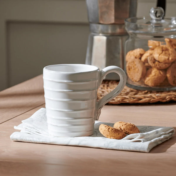 A white ceramic mug with horizontal ridges sits on a light gray cloth on a wooden table, with two small cookies beside it and a blurred coffee maker and cookie jar in the background.