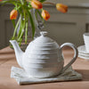 A white ceramic teapot with a ribbed texture and small beaded trim, resting on a folded cream napkin on a wooden table. A glass vase with orange tulips is blurred in the background.