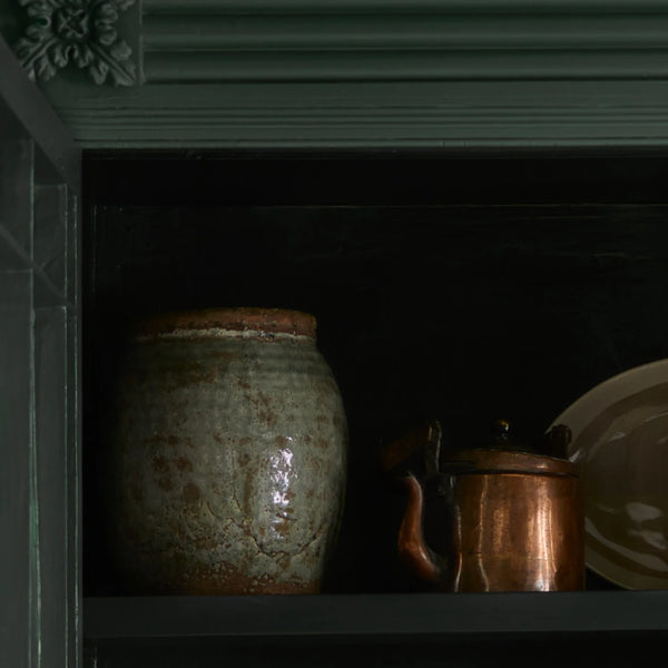 A rustic ceramic jar with a textured, earthy green and brown glaze sits on a dark shelf next to a copper-colored metal pot and a white plate, set against a dark background.