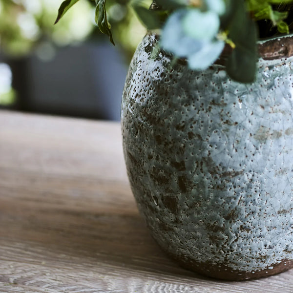 A close-up of a rustic ceramic planter with a textured, speckled blue-gray glaze and brown clay edges, holding green foliage on a light wooden surface.