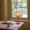 Cecile portable brass table lamp with pleated linen shade on a wooden desk in front of window overlooking a garden