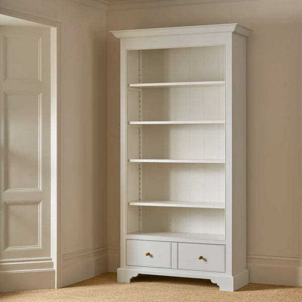 A tall white bookcase with five open shelves and two small drawers at the bottom, shown in a beige room corner next to a paneled door.