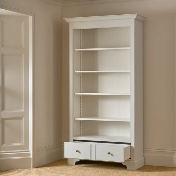 A tall white corner bookcase with five open shelves and two small drawers at the bottom, shown in a beige room corner with one drawer pulled open.