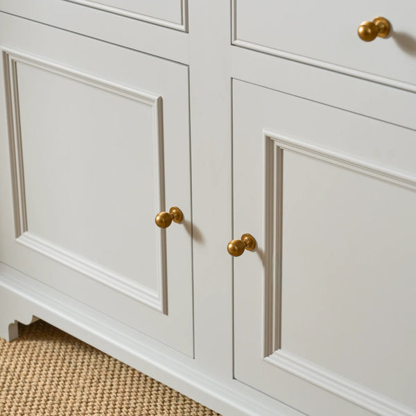 Close-up of a white paneled cabinet with two doors and round gold knobs, shown against a neutral indoor setting with a woven rug visible at the bottom.