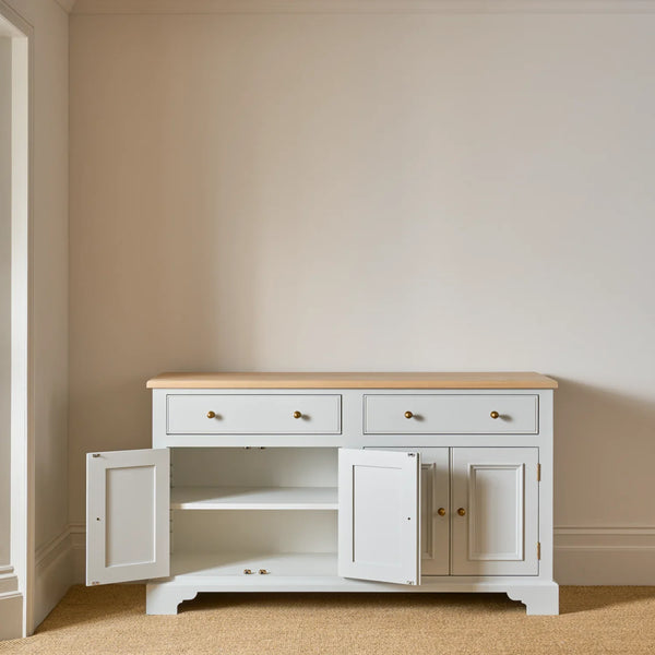 A white wooden sideboard cabinet with a light natural wood top, featuring two drawers on top, two lower cabinet doors with brass knobs, and one door swung open to reveal an interior shelf. The cabinet is set against a plain beige wall on a beige carpeted floor.