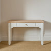 A white writing desk with a light natural wood top and a single drawer with a small brass knob, set against a neutral beige wall and textured beige carpet.