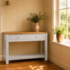 A pale grey console table with a natural oak top, styled in a warm, sunlit room with light beige panelled walls, a window, and potted plants.