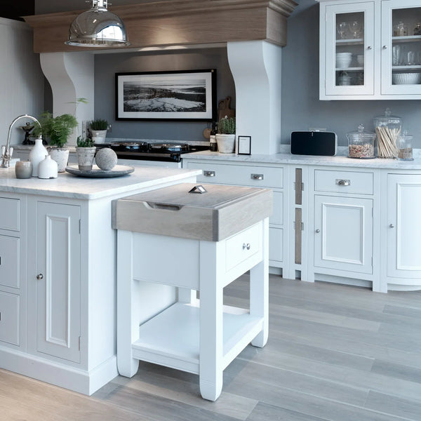A white butchers block with a natural oak end grain top with removable chopping boards. Set in a modern white kitchen setting with natural stone work surfaces and a grey oak flooring.