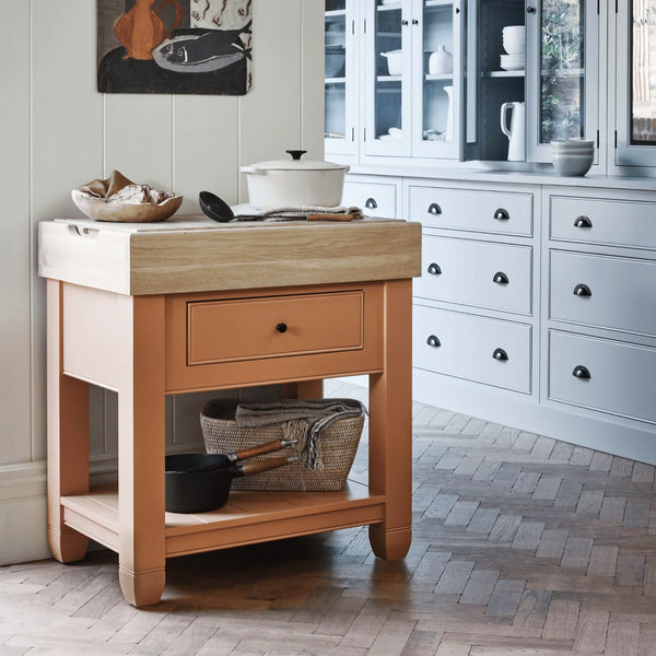 A wooden butchers block with a light oak natural wood top, a terracotta coloured drawer front, and an open lower shelf, shown in a bright kitchen setting.