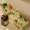 A reed diffuser bottle with amber glass and wooden reeds sits beside its illustrated product box on a beige fabric surface. The box reads Clementine & Sandalwood Diffuser and shows citrus and berry illustrations.