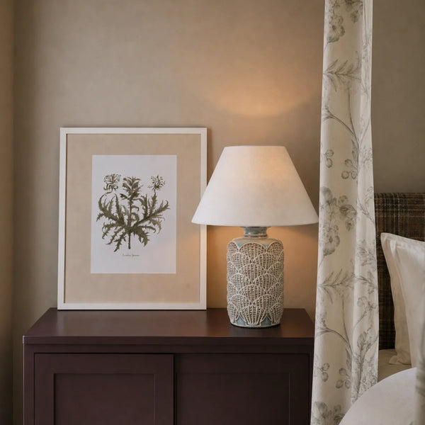 A styled bedroom scene featuring a dark wood nightstand with a lit table lamp and a framed botanical art print, set against a warm beige wall with a patterned curtain on the right.