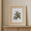 A framed botanical art print of a green plant with leaves and small flowers, displayed on a light wood console table against a beige wall.