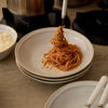 A close-up of spaghetti with red sauce being lifted from a stack of white ceramic plates on a countertop, with a bowl of grated cheese and kitchen items in the background.
