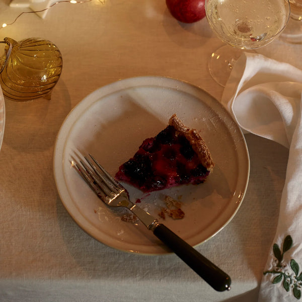 A slice of berry pie on a white ceramic plate with a fork, set on a beige tablecloth with a wine glass, napkin, and amber glass decor in the background.