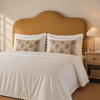 Super King mustard yellow upholstered headboard, a neatly made bed in a warm, softly lit bedroom. The bedding set features a white ruffled duvet cover and matching white pillow shams with two decorative beige pillows printed with a brown damask floral pattern.