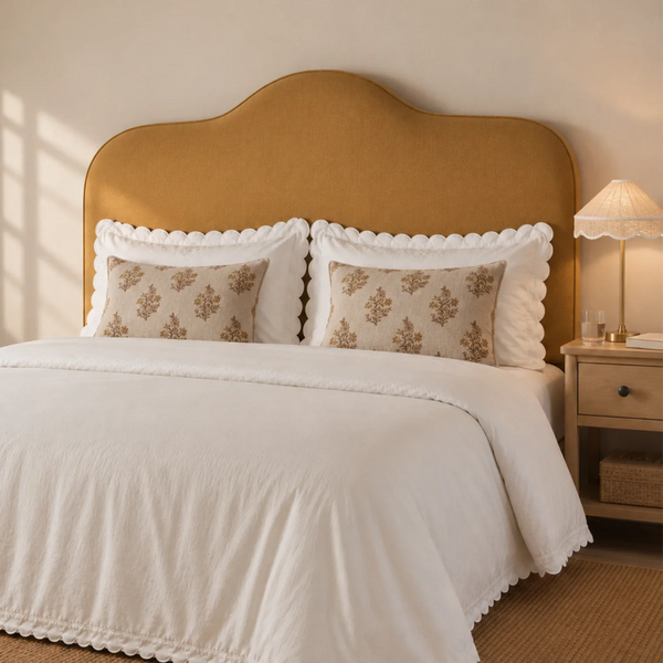 Super King mustard yellow upholstered headboard, a neatly made bed in a warm, softly lit bedroom. The bedding set features a white ruffled duvet cover and matching white pillow shams with two decorative beige pillows printed with a brown damask floral pattern.