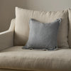 A square gray throw pillow with a textured linen-like fabric, styled on a beige upholstered armchair against a neutral beige wall.