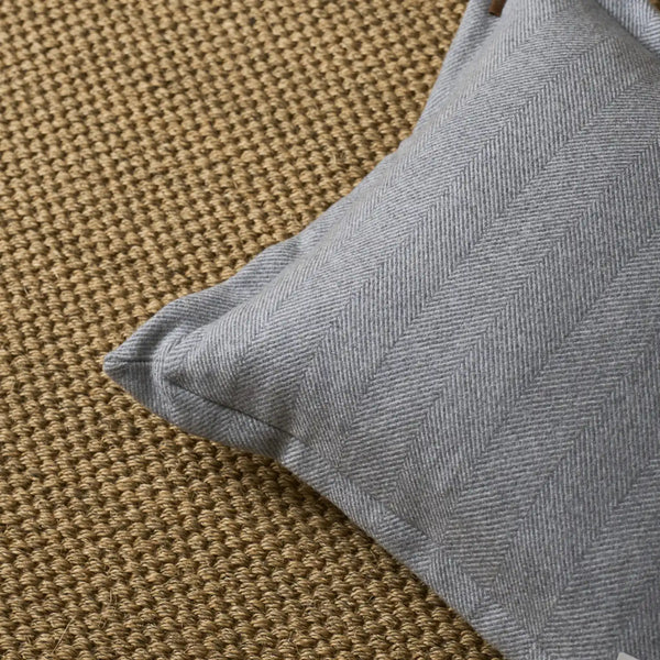 A close-up of a light gray herringbone-patterned throw pillow resting on a textured tan woven rug.