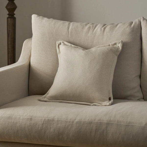 A light beige textured throw pillow resting on a matching beige upholstered armchair, with a larger matching back cushion behind it.