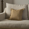 A square tan linen throw pillow with ruffled edges resting on a light beige upholstered armchair.