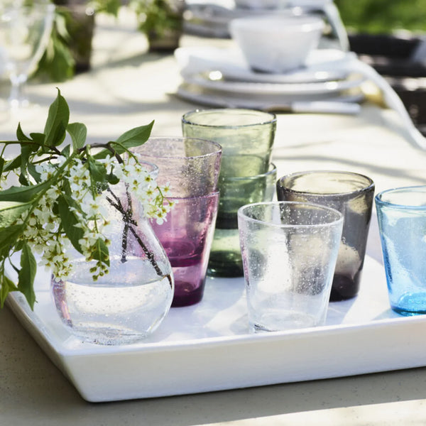 A set of colorful, textured glass tumblers and a matching glass vase arranged on a white rectangular tray outdoors on a sunlit table, with green leaves and white flowers in the foreground.