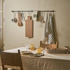 A wall-mounted black metal kitchen utensil rack hanging in a rustic dining room. The rack holds wooden cutting boards, metal pots, spoons, a whisk, and striped towels, above a table set with glasses and plates.