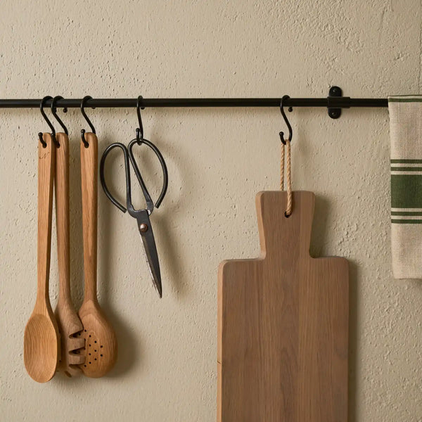 A black metal wall-mounted hanging rod with multiple kitchen utensils and a wooden cutting board suspended from black hooks against a textured beige wall.