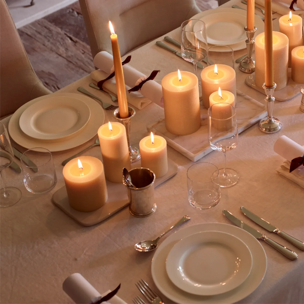 A warmly lit dining table set with cream candles in various heights, white plates, silver cutlery, and clear glassware in a softly lit indoor setting.