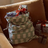 A large woven holiday gift sack with a green and cream geometric pattern sits on a brown velvet armchair, filled with wrapped presents and a small ornament. Two additional wrapped gifts with red and dark ribbons rest beside it.
