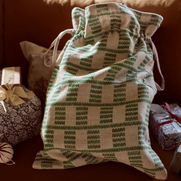 A knitted Christmas stocking bag with a green and cream geometric pattern, styled on a brown leather sofa next to wrapped gifts.