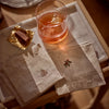 Two beige linen napkins with delicate embroidered holly motifs, arranged on a marble tray next to a glass of amber drink and a wrapped chocolate.