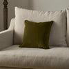 An olive green velvet square throw pillow resting on a light beige upholstered armchair.