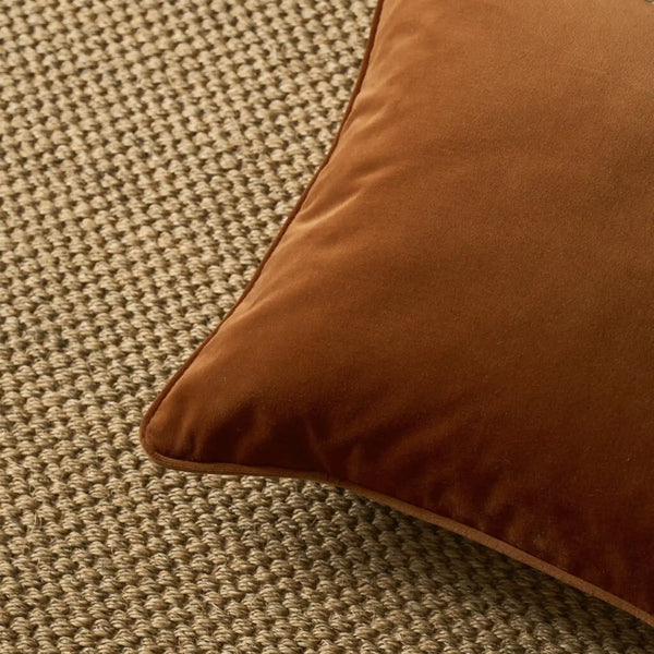 A close-up of a square brown velvet throw pillow resting on a textured beige woven rug.