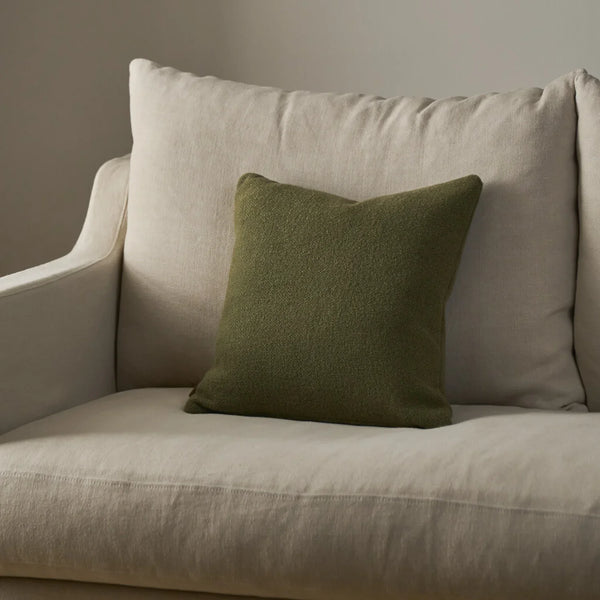 An olive green textured throw pillow resting on a light beige upholstered armchair against a neutral gray wall.